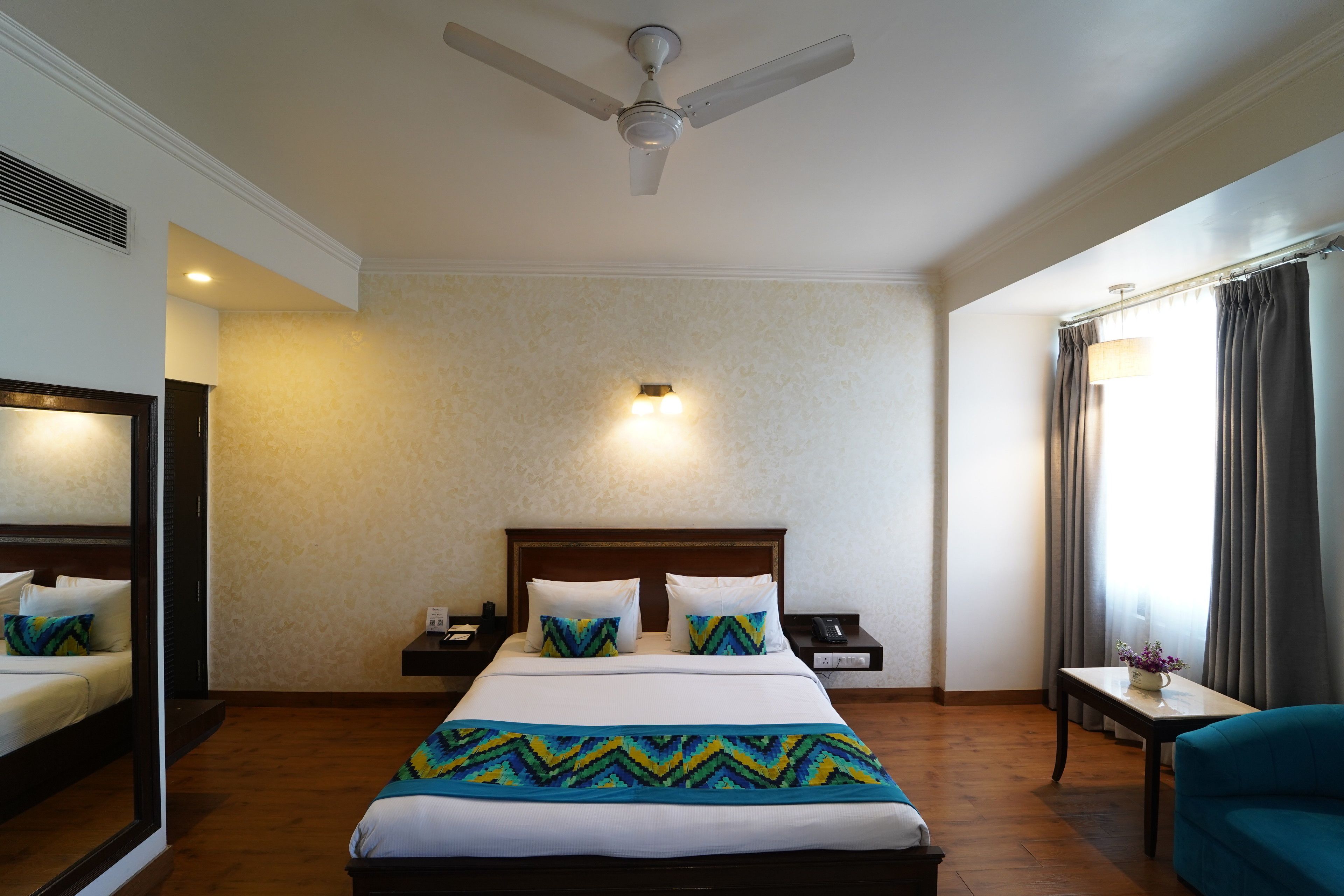 A hotel room with a large bed, a ceiling fan, a bedside table with a lamp, a nightstand, and a window with curtains.