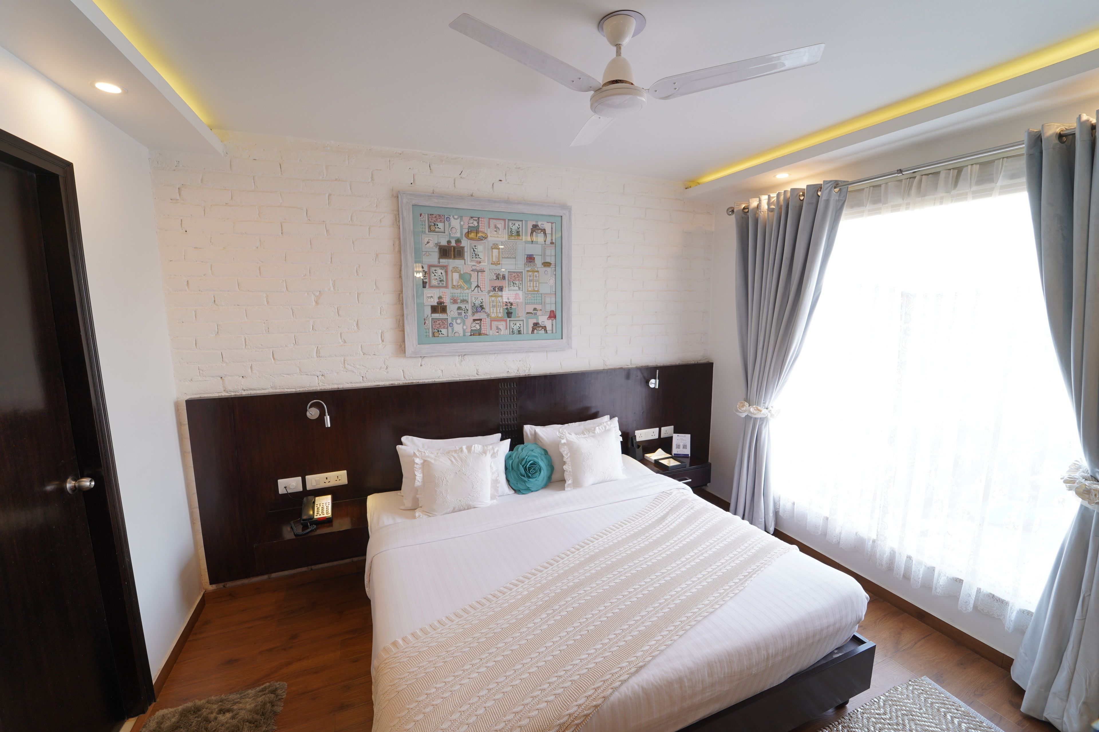 A hotel room with a large bed, a ceiling fan, a white brick wall, a framed picture, and a window with curtains.