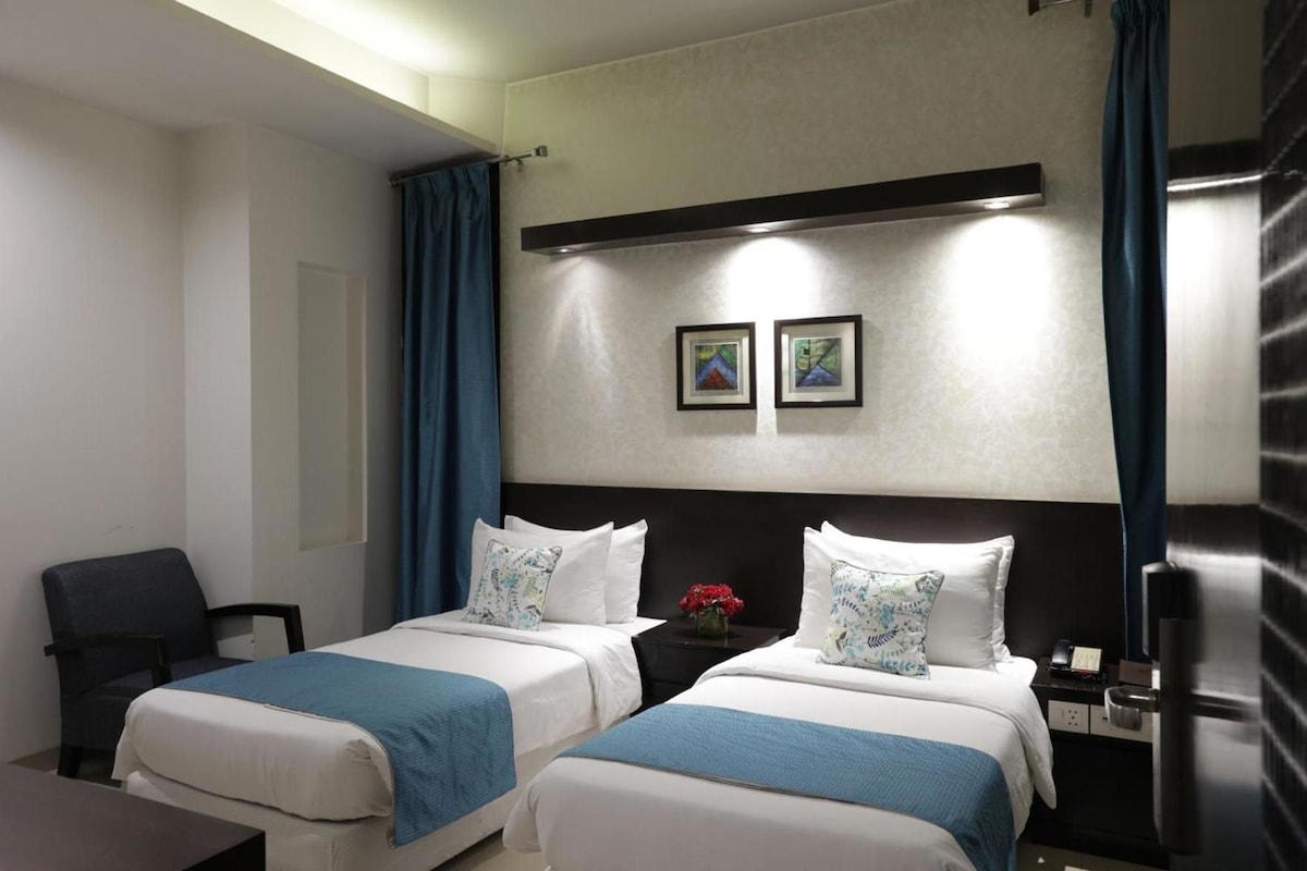 Habitación de hotel con dos camas, un escritorio, una silla, una mesita de noche y una lámpara de pared.