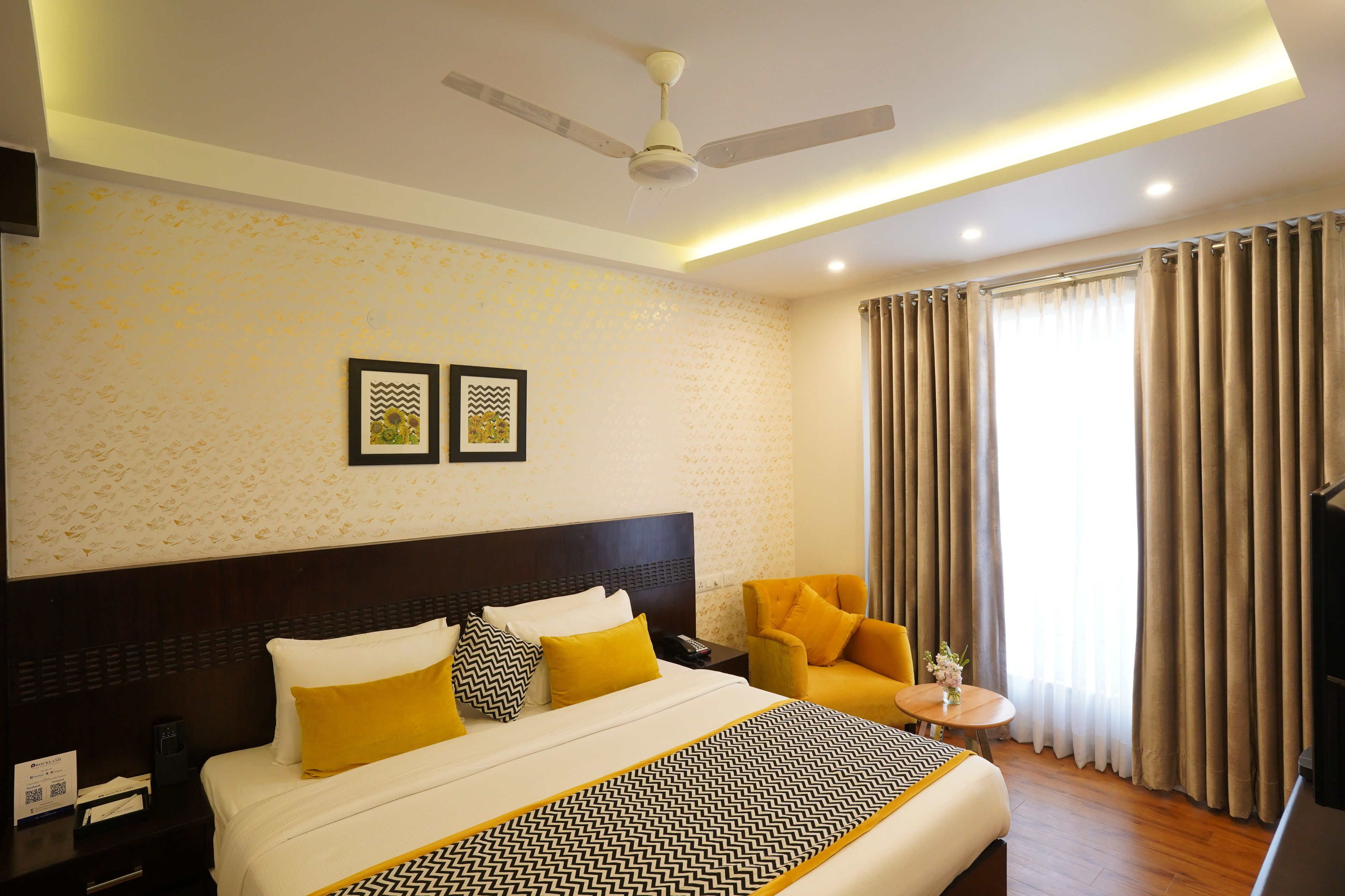 A hotel room with a large bed, a nightstand, a chair, a small table, a ceiling fan, and a window with curtains.