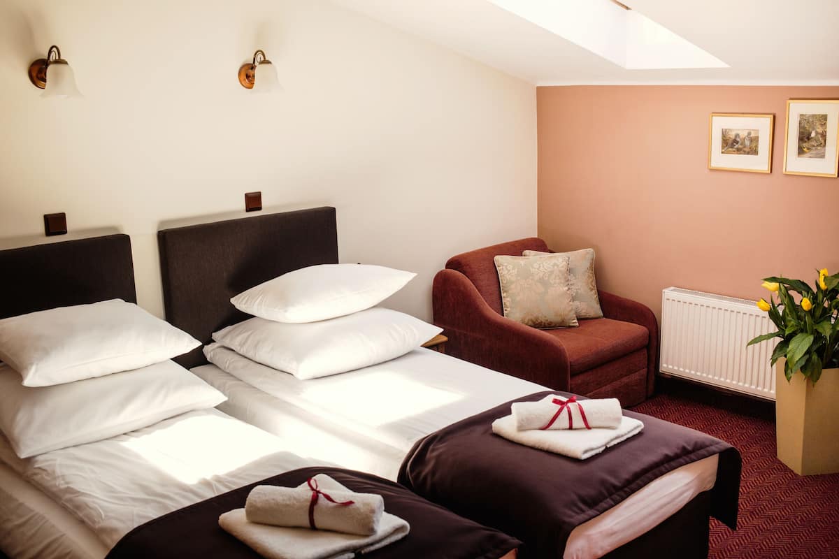 A hotel room with two beds, a red armchair, a radiator, and a window with natural light.