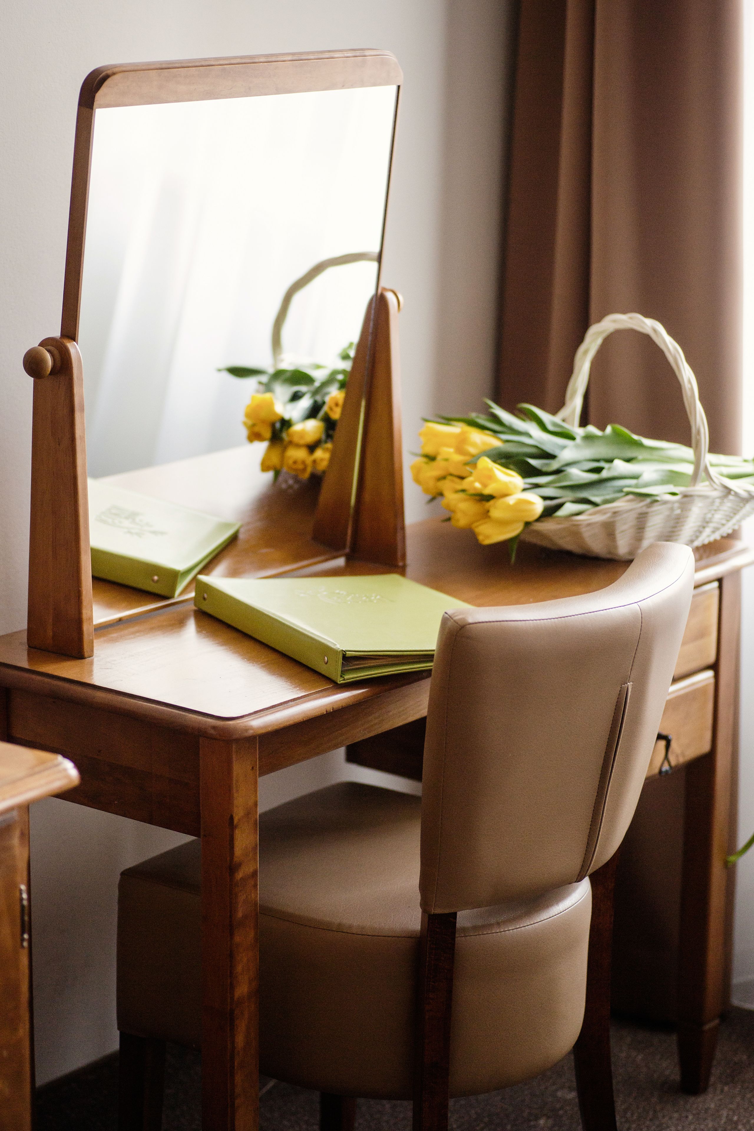 Un bureau en bois avec un miroir, un carnet vert et un panier de fleurs jaunes.