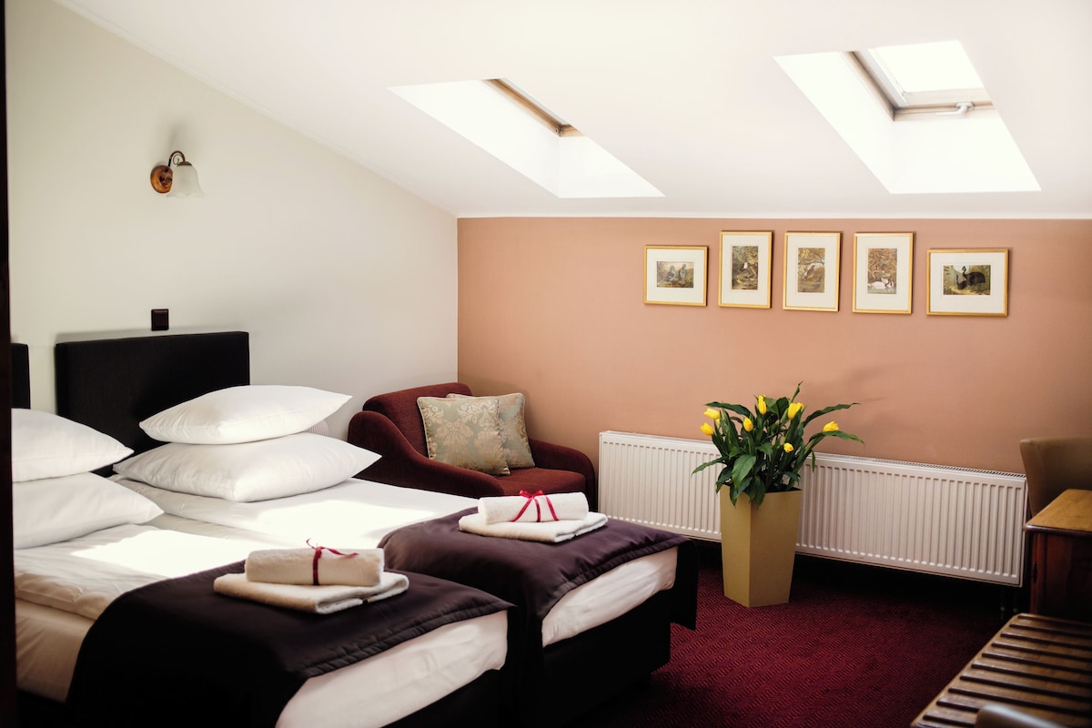 A hotel room with a bed, a red armchair, a small table, and a radiator. There are four framed pictures on the wall and a potted plant.
