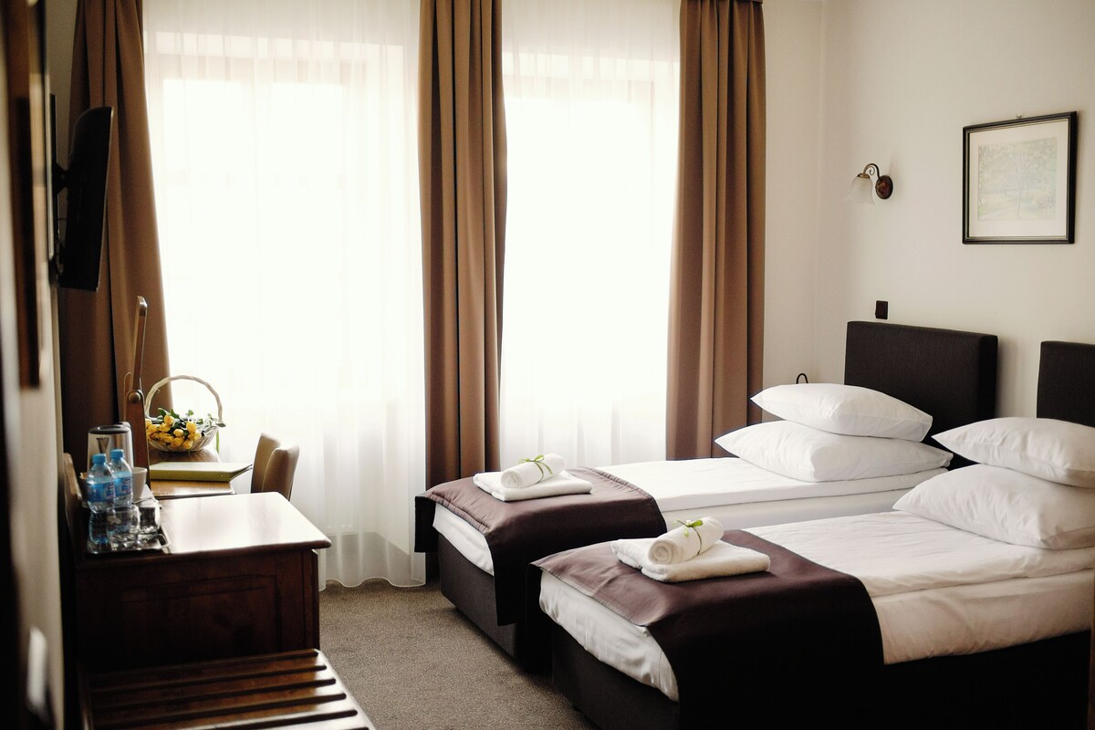 A hotel room with two beds, a desk with a chair, a small table with a bowl of fruit, and a framed picture on the wall.