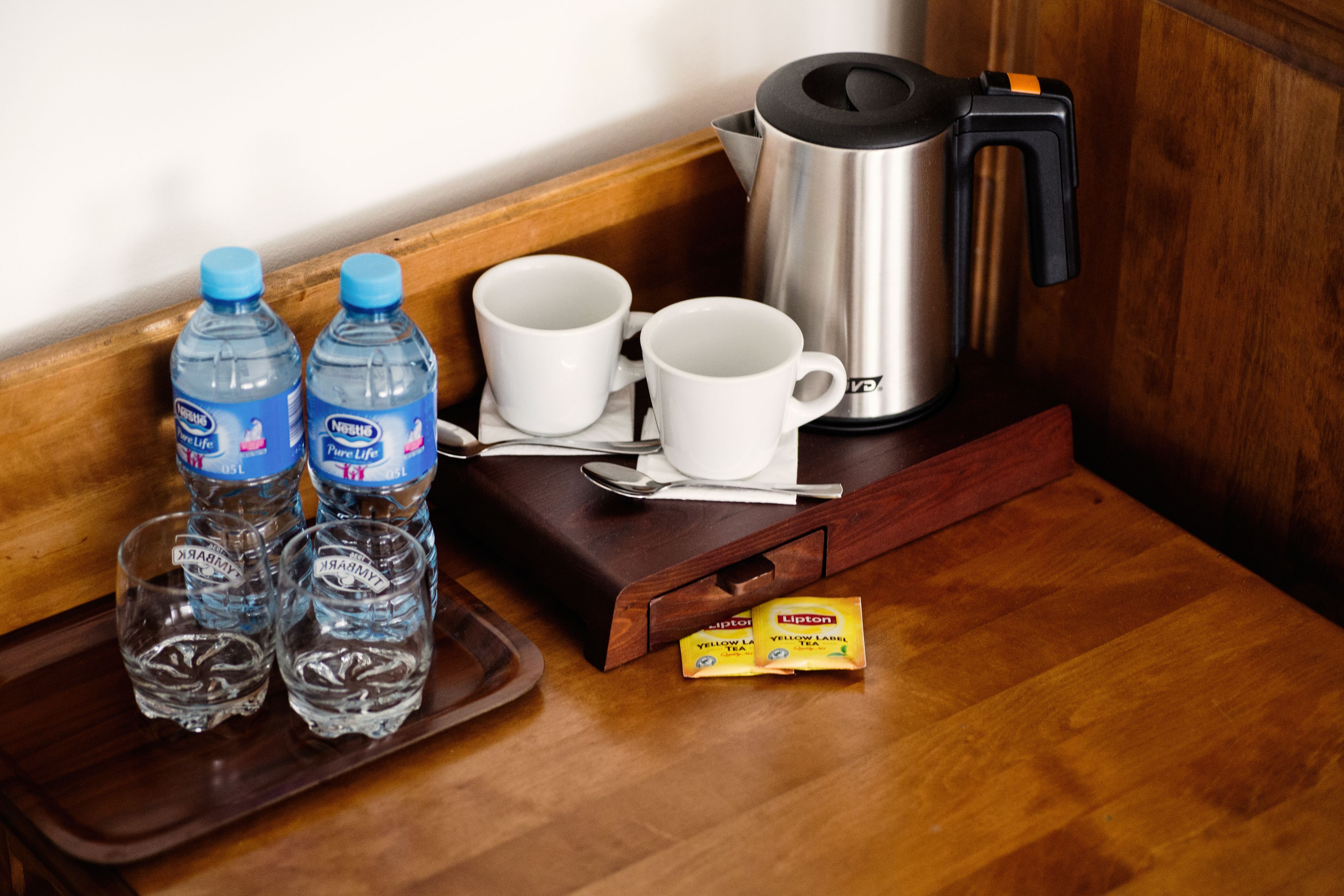 Una bandeja de madera con botellas de agua, vasos blancos, una cuchara, un sobre de té Lipton y un hervidor de acero inoxidable sobre una mesa de madera.