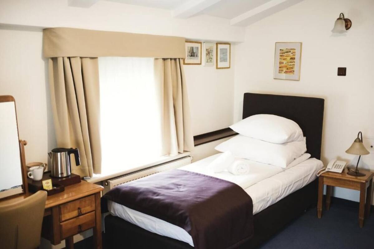 A hotel room with a bed, a bedside table with a lamp, a small desk with a kettle, and a window with curtains.