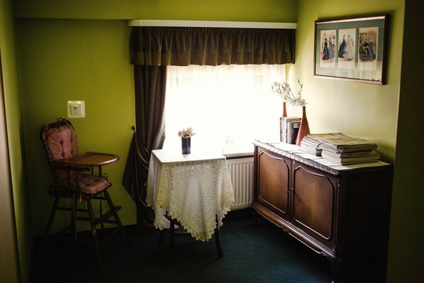 A room with a green wall, a radiator, a table with a lace tablecloth, a chair, a cabinet with books, and a window with curtains.