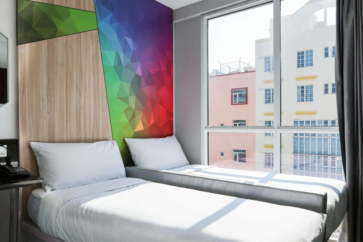 A hotel room with a bed, a bedside table, and a window with a view of buildings.