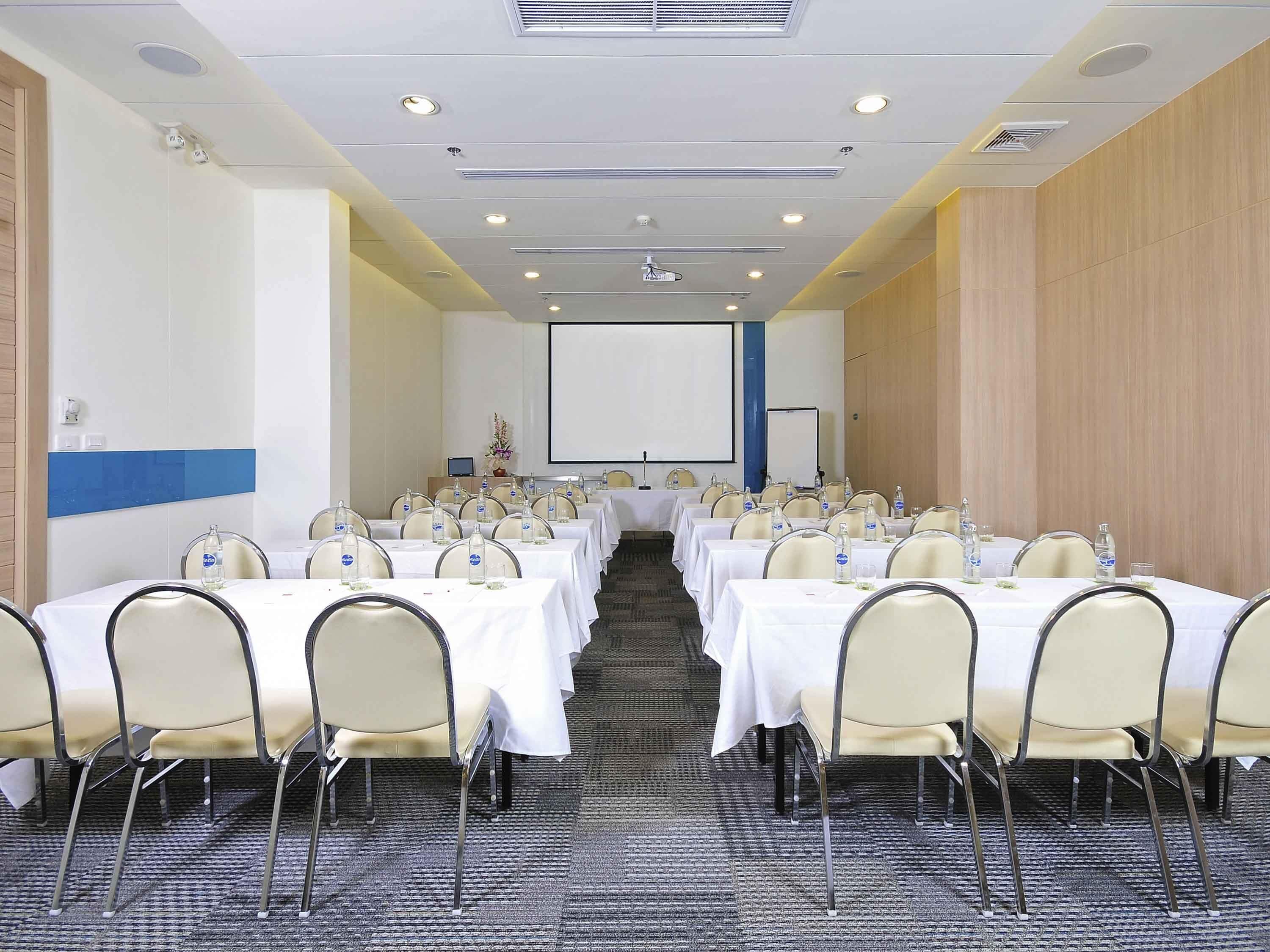 A conference room with multiple tables, chairs, a projector screen, and a carpeted floor.