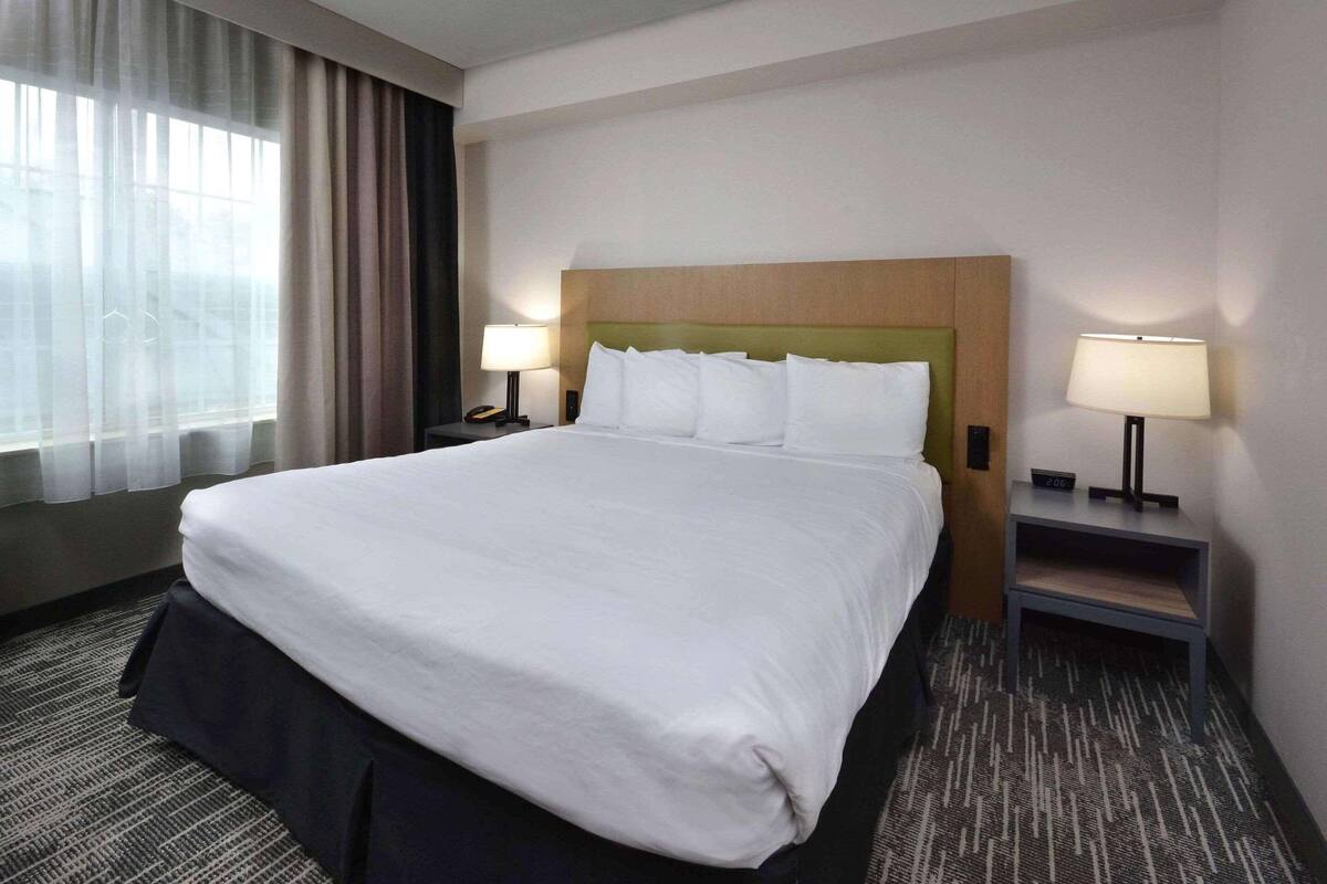 A hotel room with a large bed, two bedside tables with lamps, a window with curtains, and a carpeted floor.