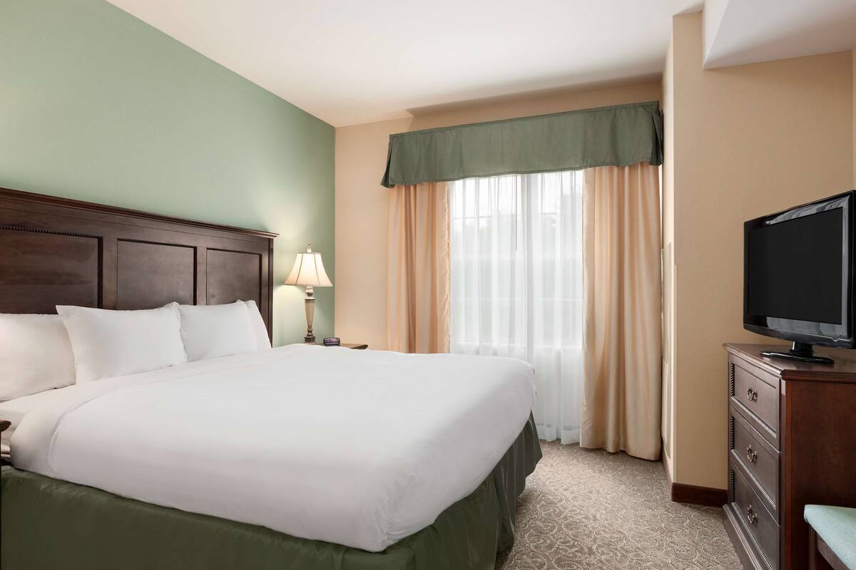 A hotel room with a large bed, a television on a dresser, a lamp, and a window with curtains.