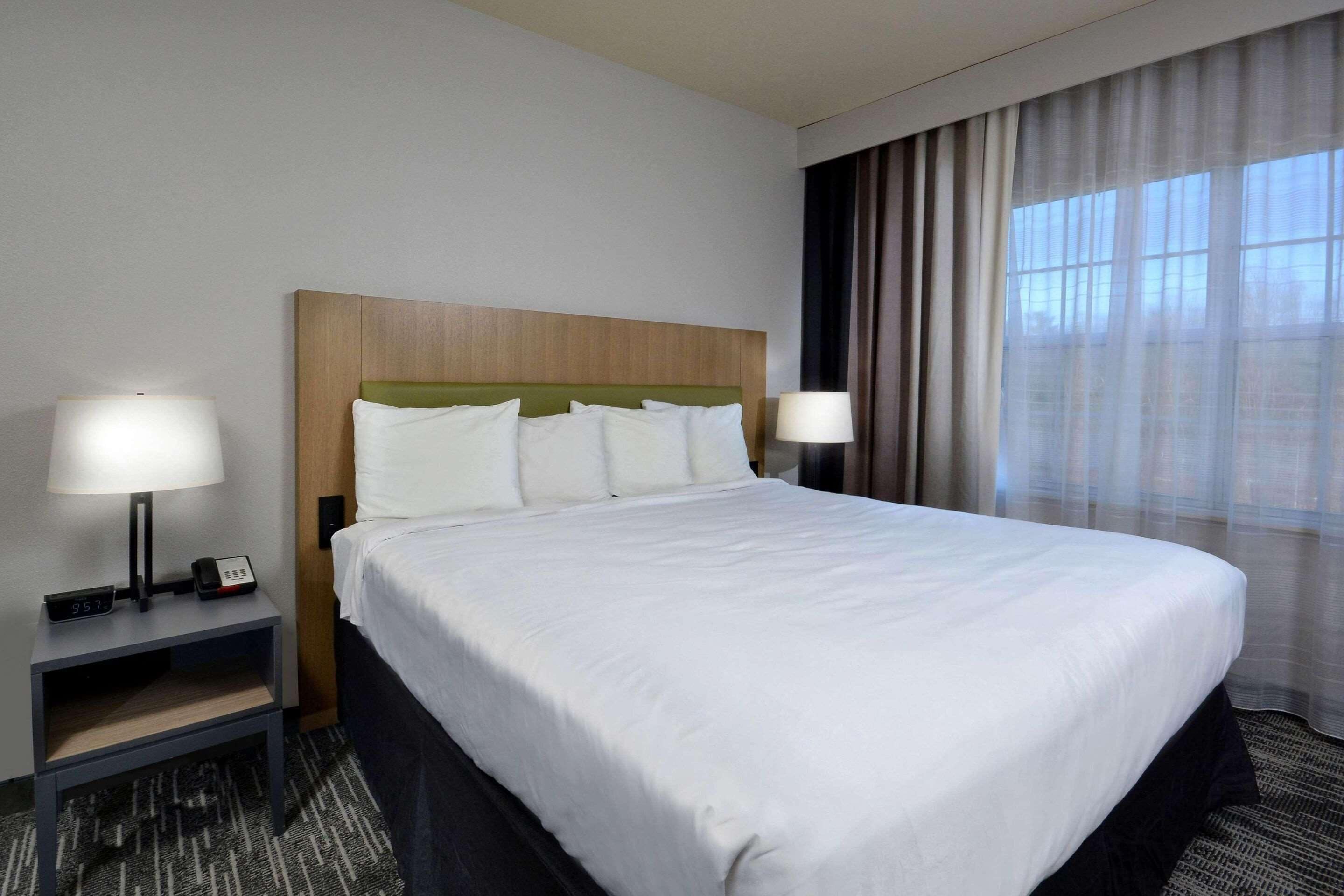 Une chambre d’hôtel avec un grand lit, deux tables de chevet avec des lampes, une tête de lit en bois et une vue sur la ville par la fenêtre.