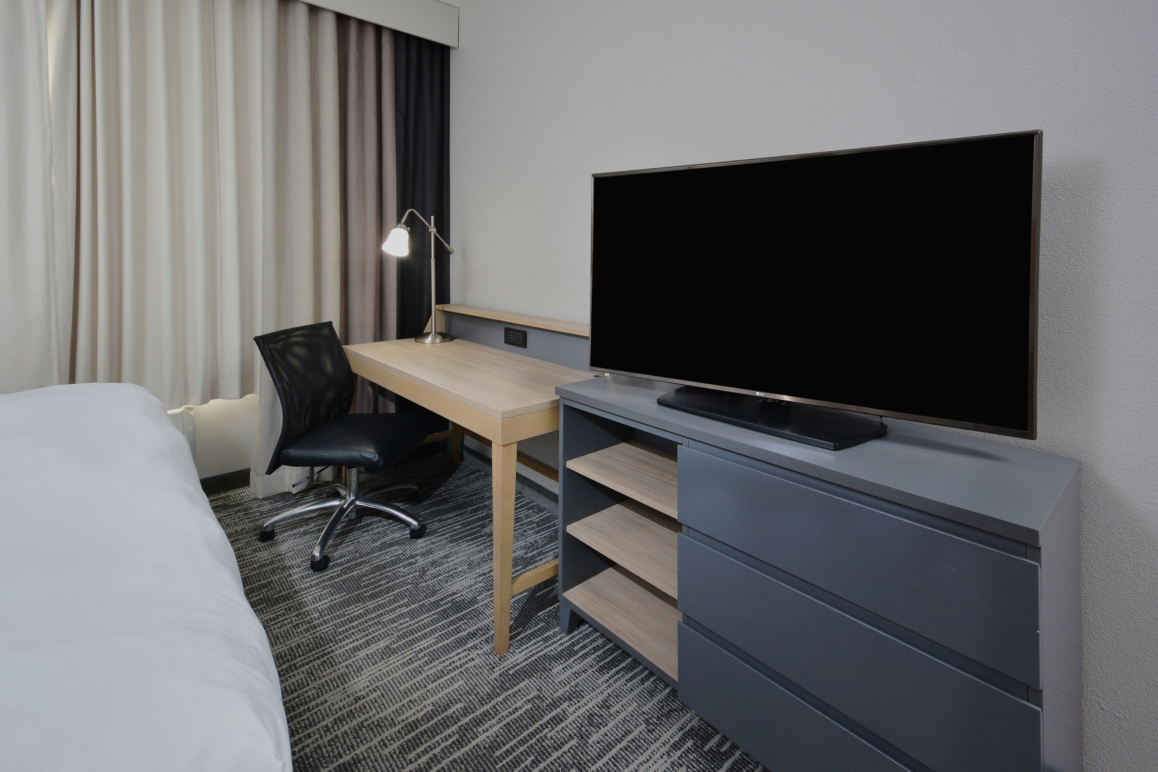 A hotel room with a bed, a desk with a chair, a television, and a desk lamp.