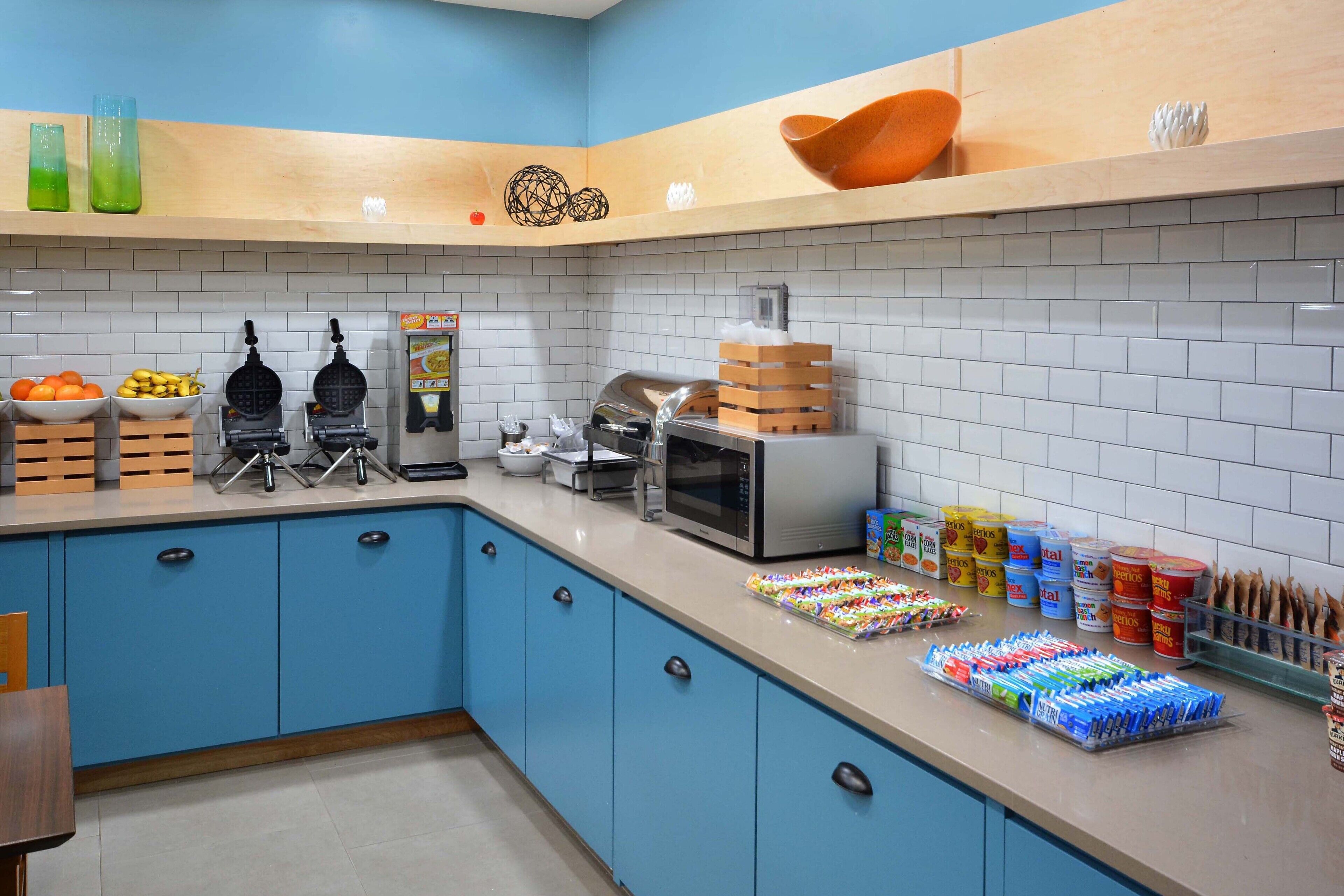 A kitchen with blue cabinets, a microwave, waffle maker, and various food items on the counter.