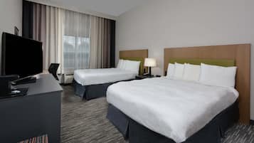A hotel room with two beds, a desk with a flat-screen TV, and a large window with curtains.