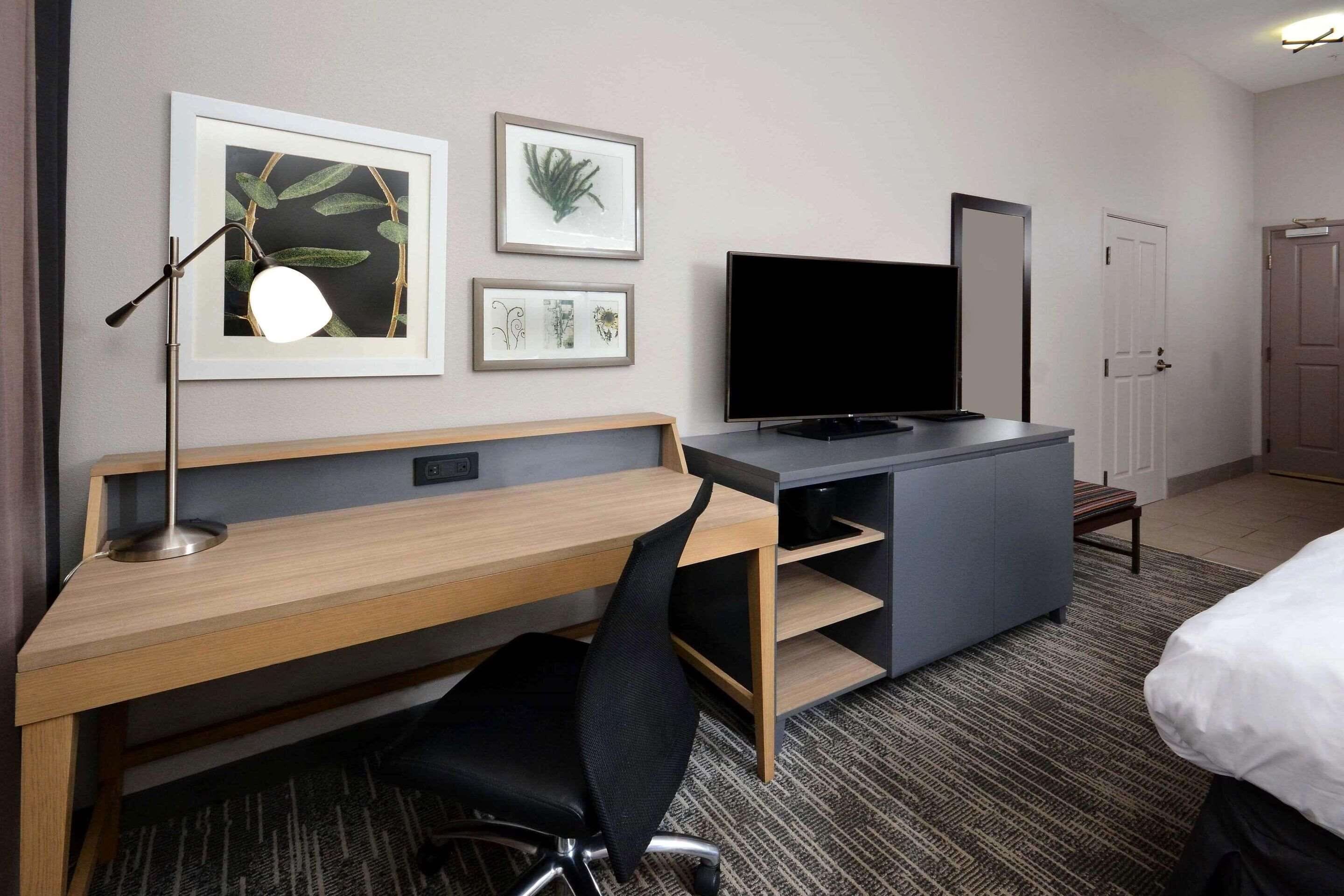 Una habitación de hotel con un escritorio de madera, una silla de oficina negra, un televisor de pantalla plana y una cama.