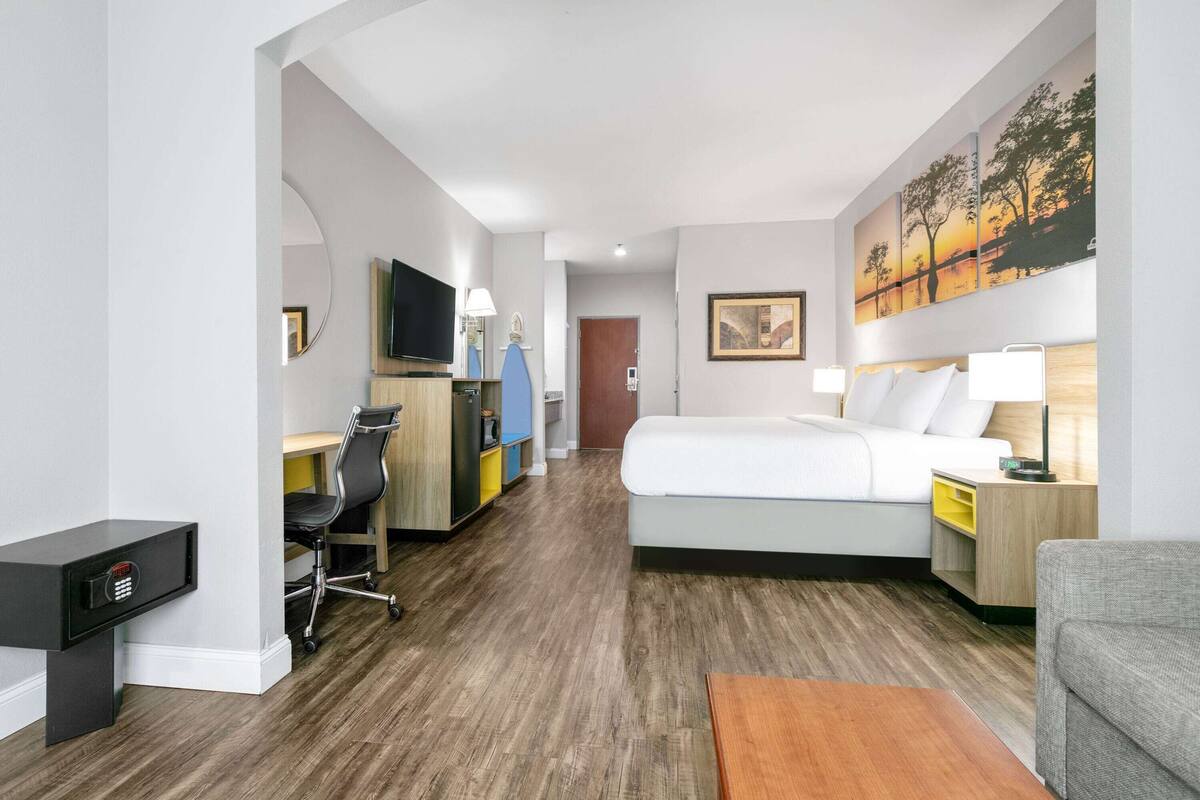 A hotel room with a bed, desk, chair, TV, and artwork on the wall.