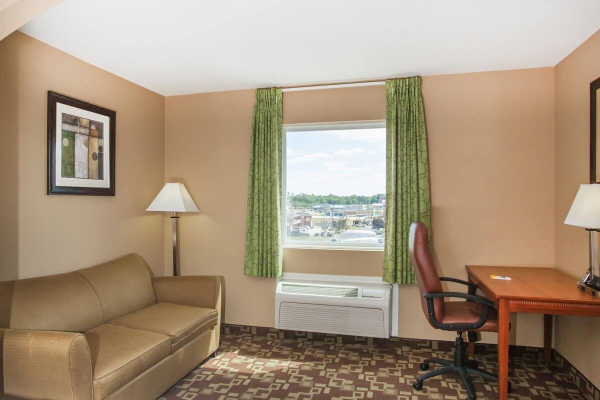 A hotel room with a sofa, a desk, a chair, a window with curtains, an air conditioner, and a framed picture on the wall.
