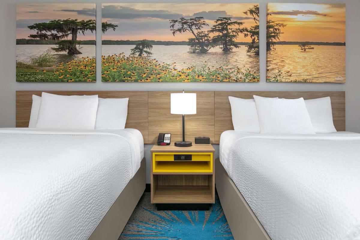 A hotel room with two beds, a nightstand with a lamp, and a wall-mounted painting of a sunset over water.