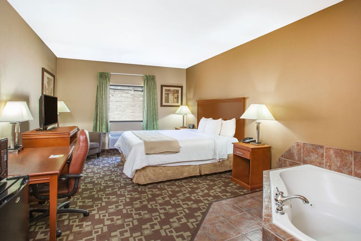 A hotel room with a bed, desk, chair, TV, lamps, and a bathtub.