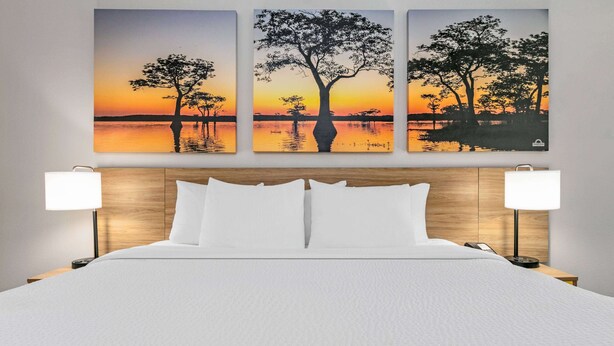 A hotel room with a large bed, two bedside lamps, and three paintings of a sunset over a lake. - Days Inn & Suites by Wyndham Cabot (Cabot)