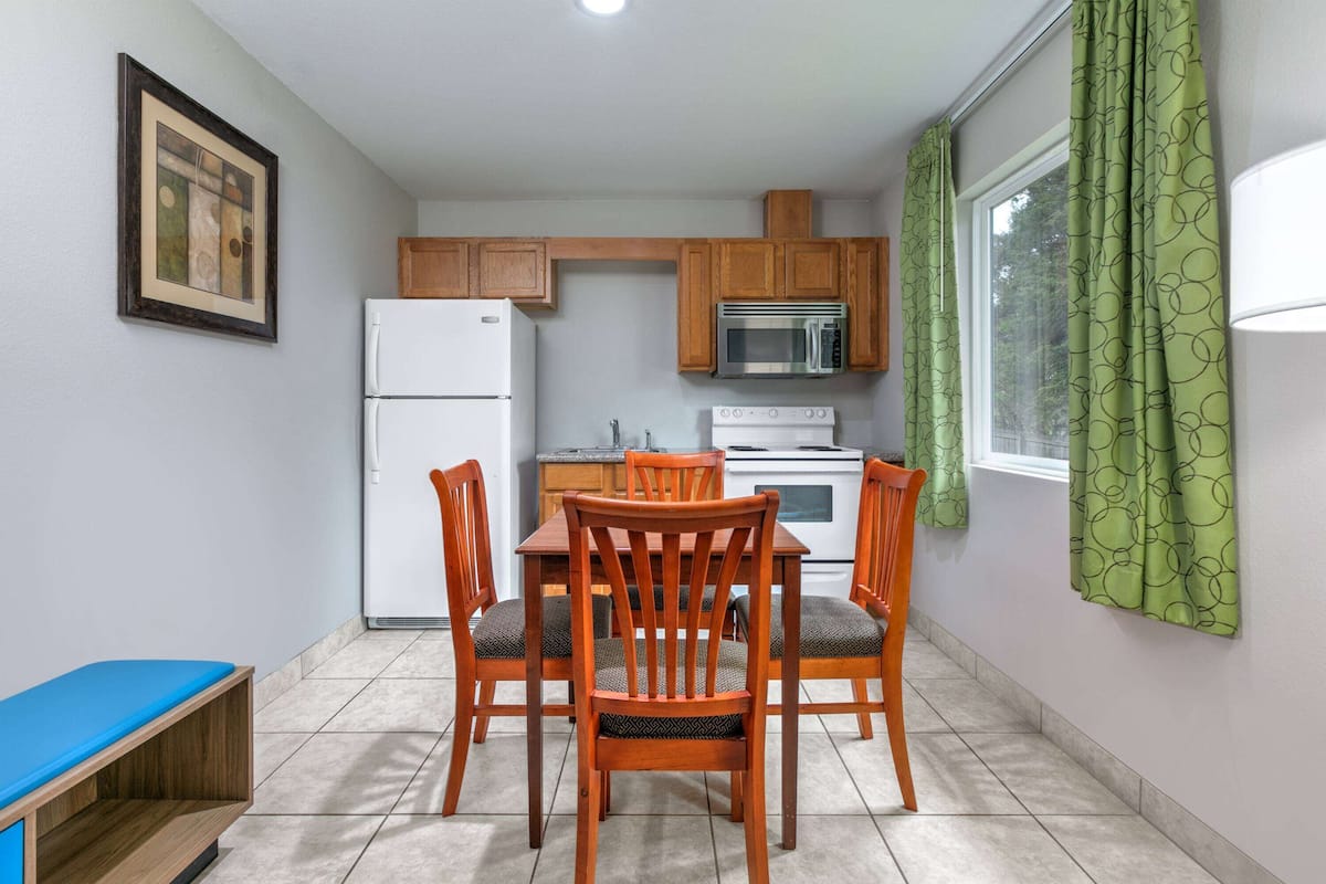 A compact kitchen with wooden cabinets, a white refrigerator, a microwave, and a stove. There is a dining table with four chairs, and a window with green curtains.