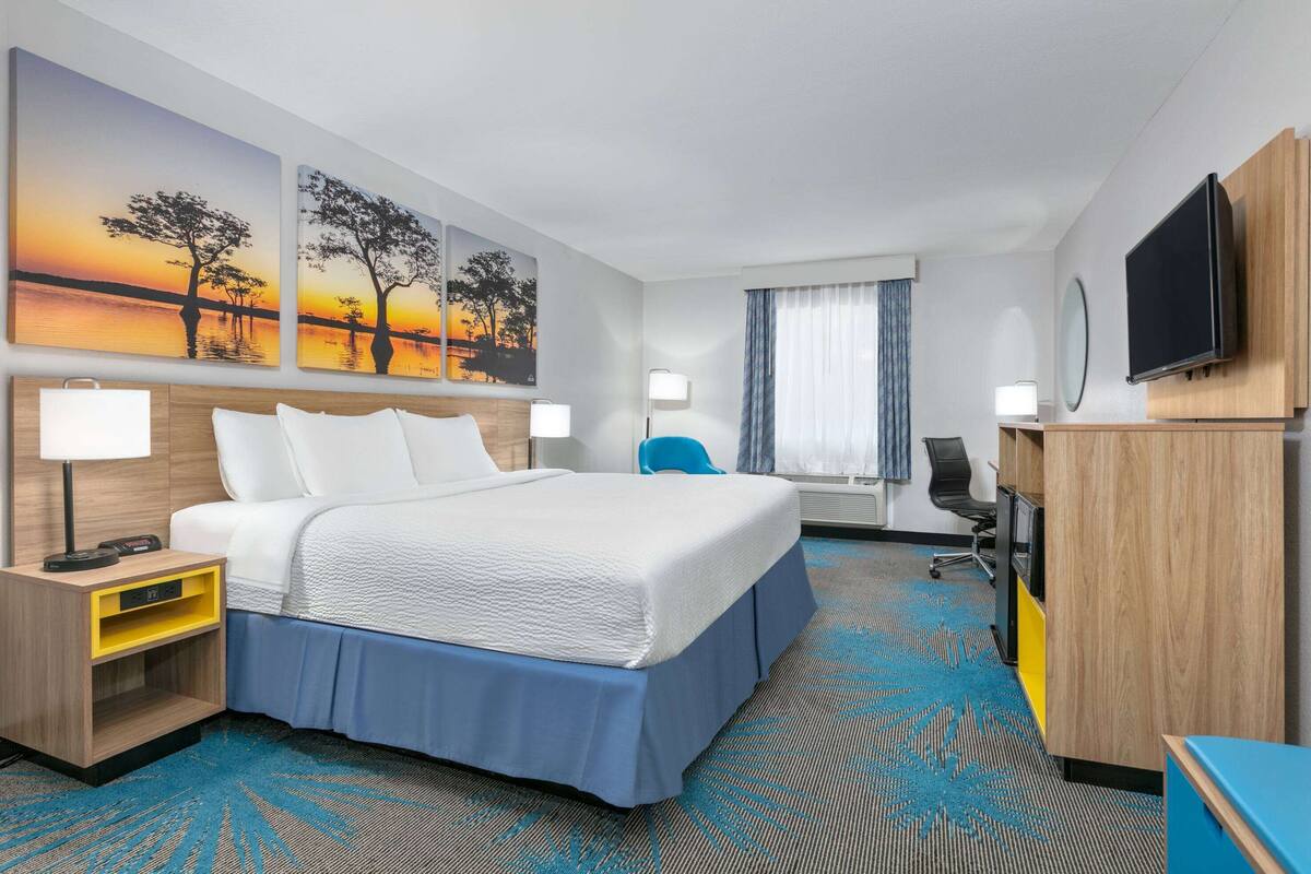 A hotel room with a large bed, a desk, a chair, a TV, and artwork on the walls.