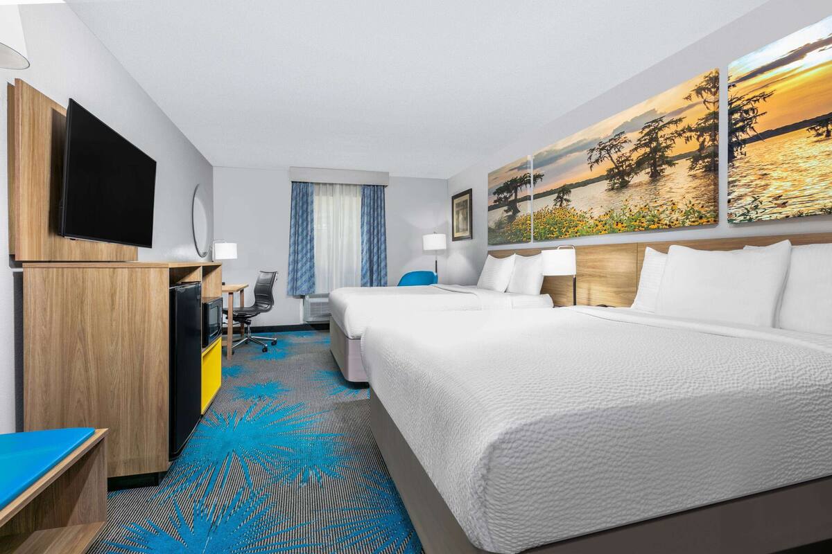 A hotel room with a large bed, a desk, a television, and a painting of a sunset.