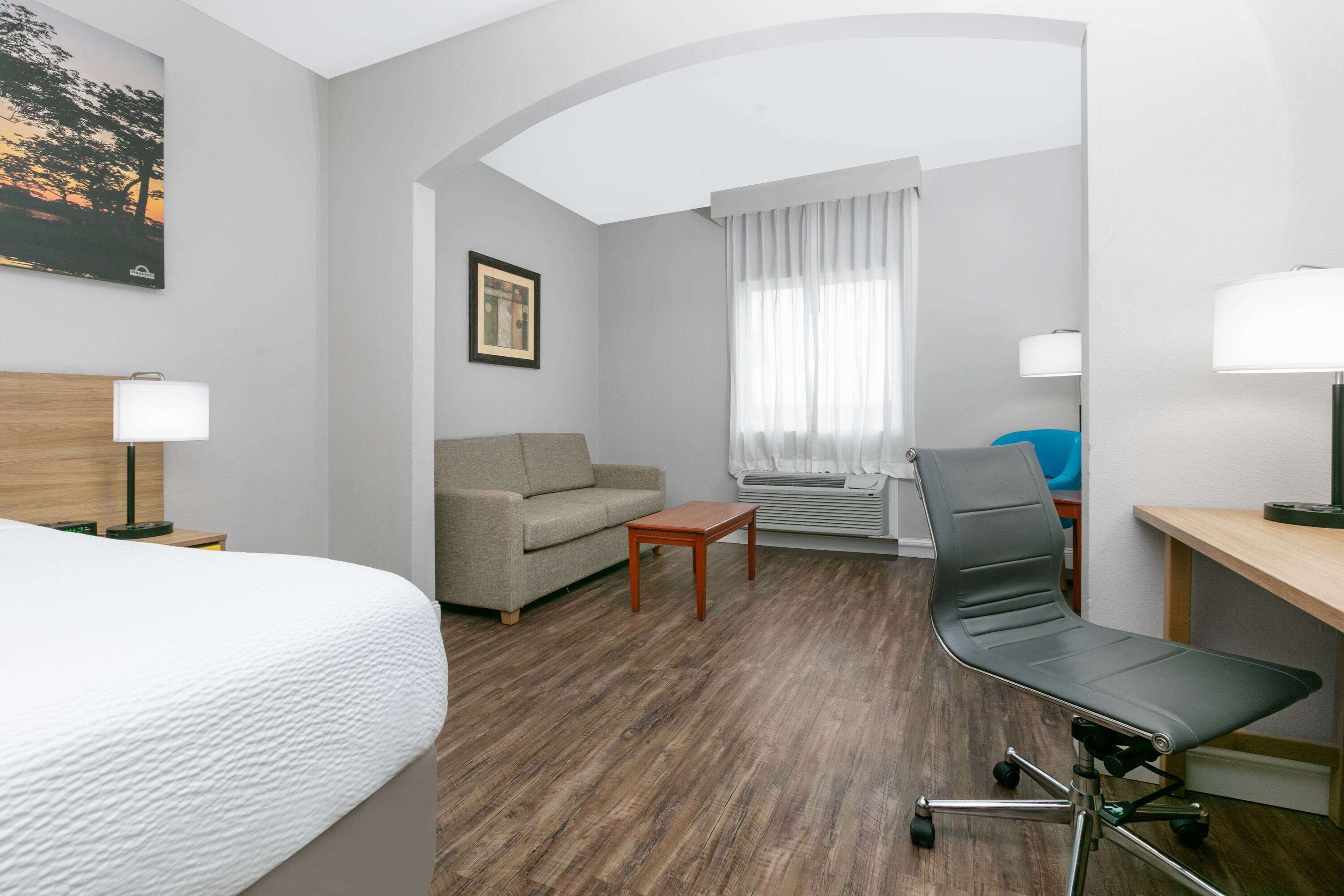 Habitación de hotel con una cama, un sofá gris, una mesa de centro de madera, un escritorio con una silla y una ventana con cortinas.