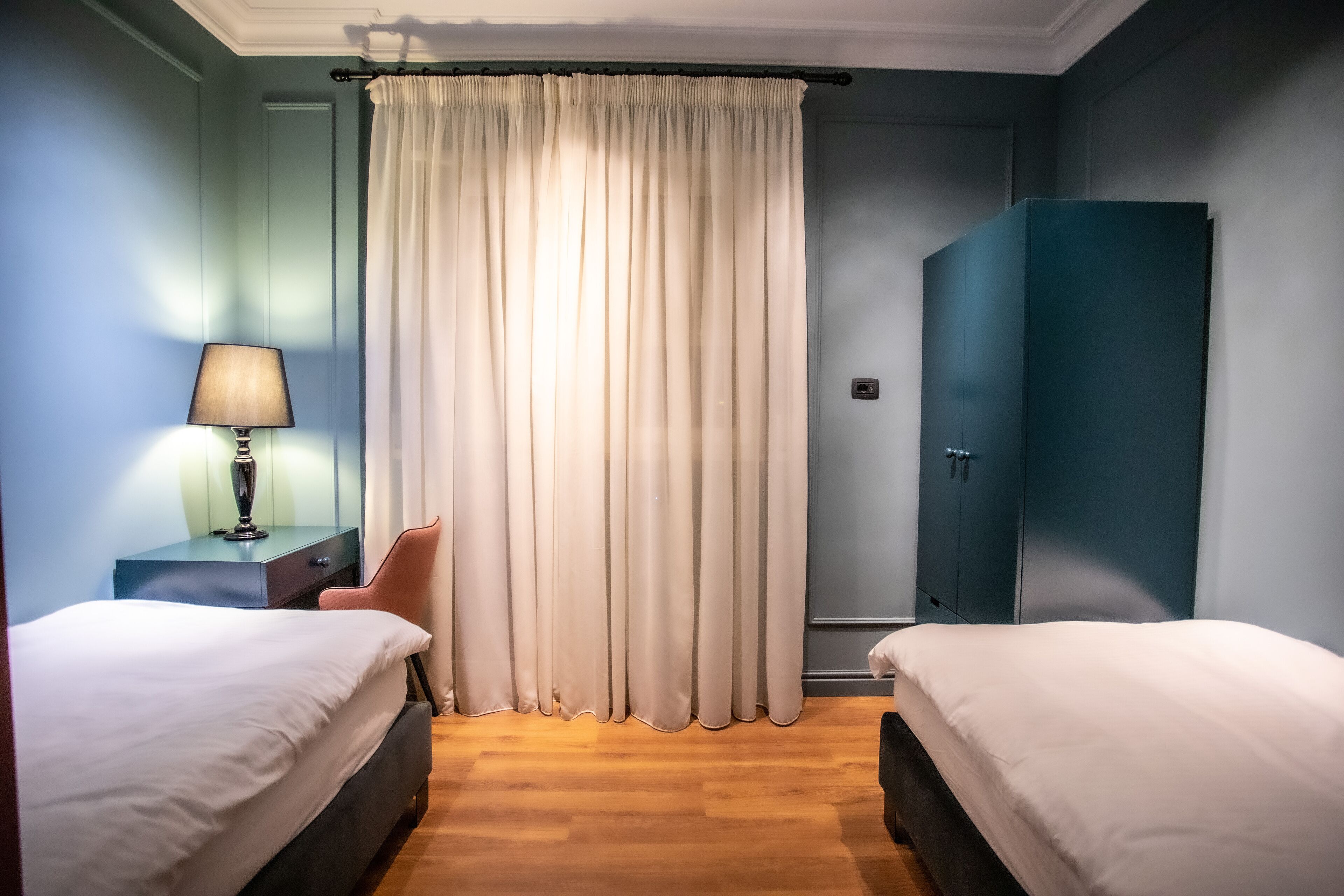 Quarto de hotel com duas camas, uma escrivaninha com luminária, um guarda-roupa e uma cortina.