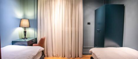 Quarto de hotel com duas camas, uma escrivaninha com luminária, um guarda-roupa e uma cortina.