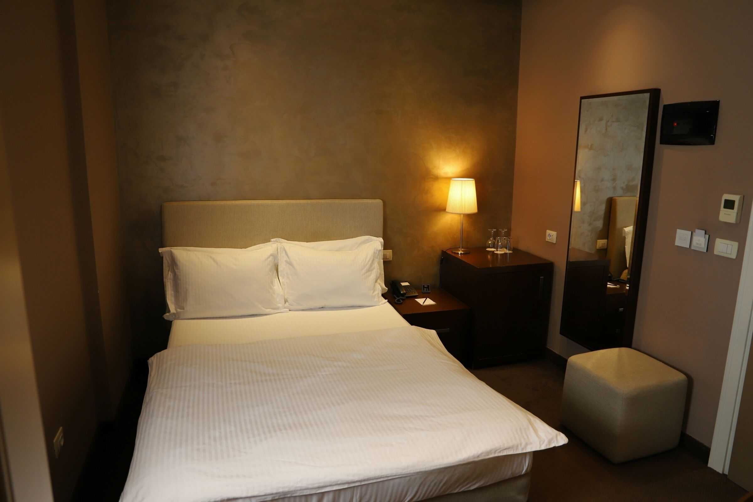 Quarto de hotel com cama, luminária de cabeceira, mesa de cabeceira, espelho e um pequeno banquinho.