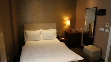 Quarto de hotel com cama, luminária de cabeceira, mesa de cabeceira, espelho e um pequeno banquinho.