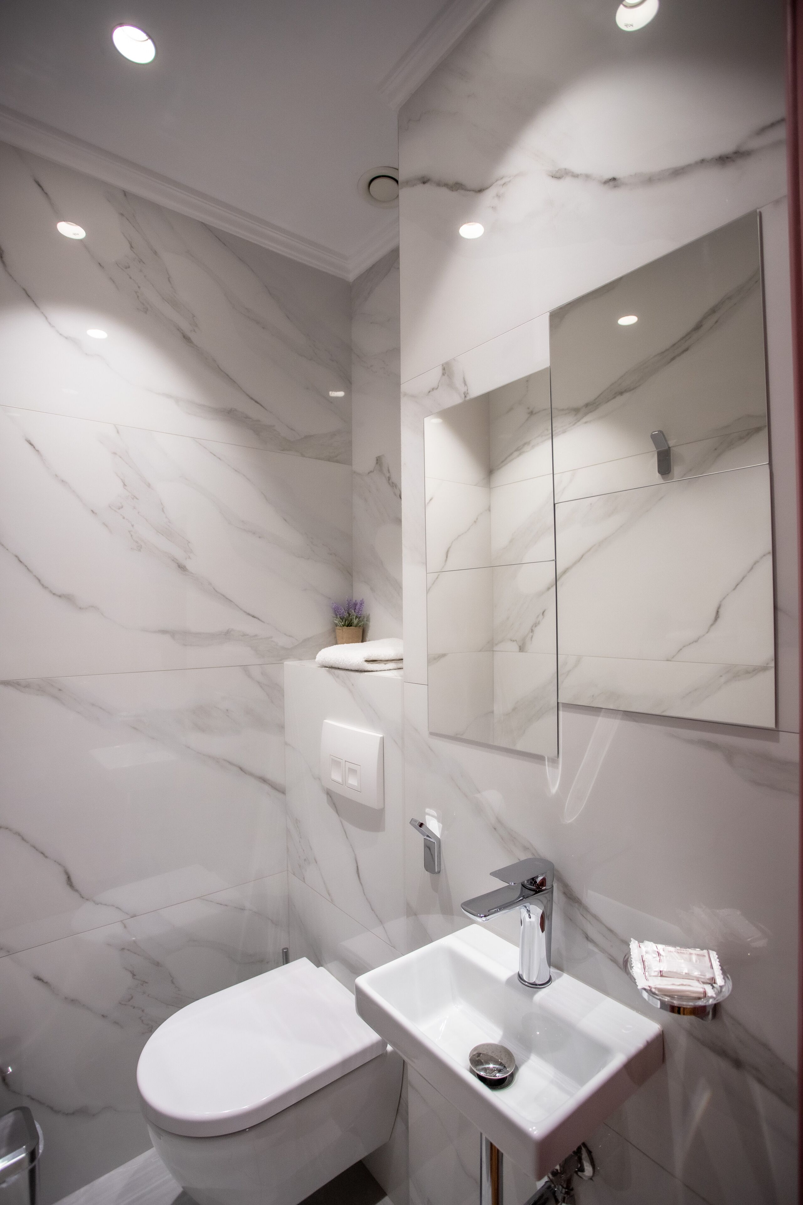 Un baño moderno con pared de mármol blanco, un espejo grande, un lavabo blanco y un inodoro.