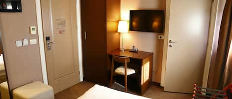 A hotel room with a bed, a desk with a chair, a TV mounted on the wall, and a door.