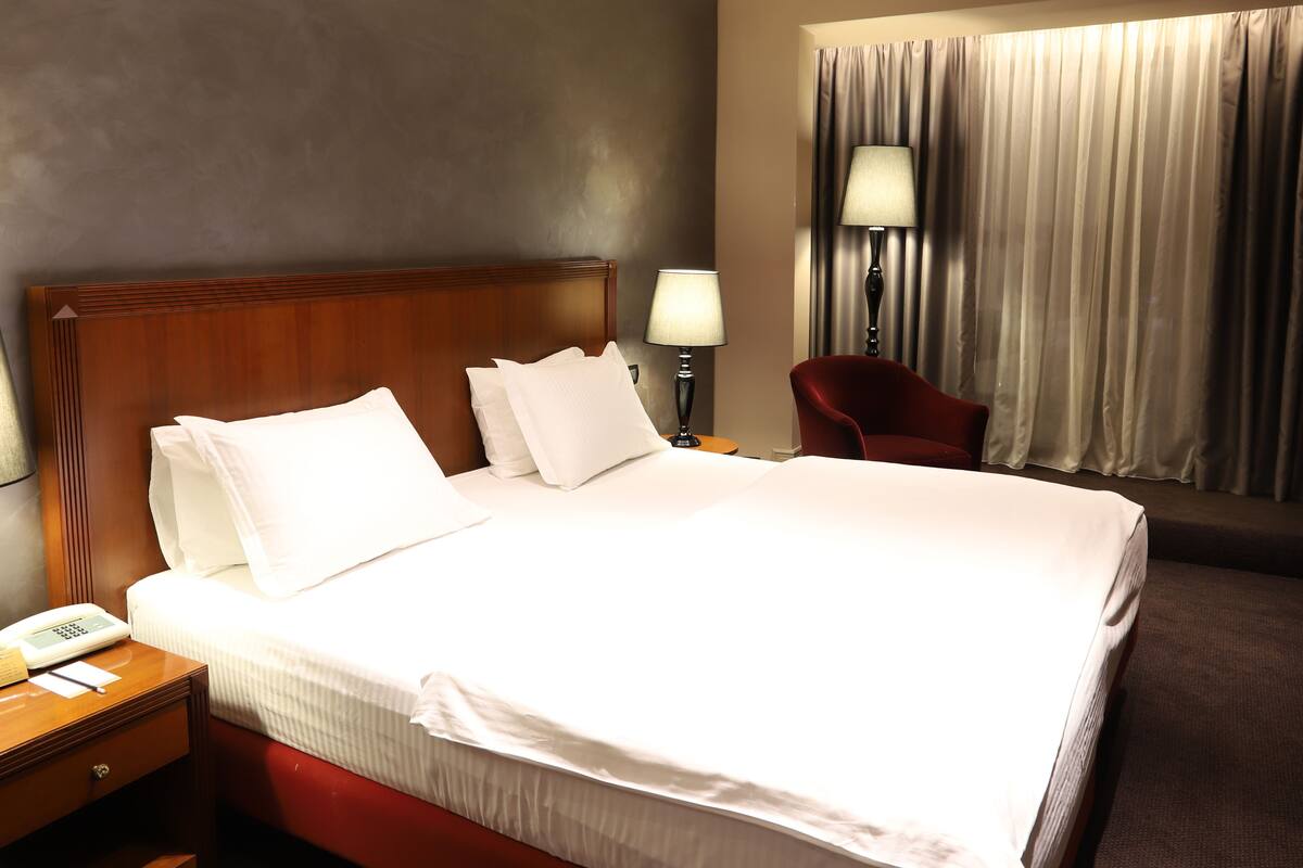 A hotel room with a large bed, a nightstand with a phone and a lamp, a red chair, and a window with curtains.