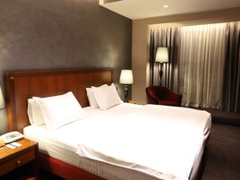 A hotel room with a large bed, a nightstand with a phone and a lamp, a red chair, and a window with curtains.