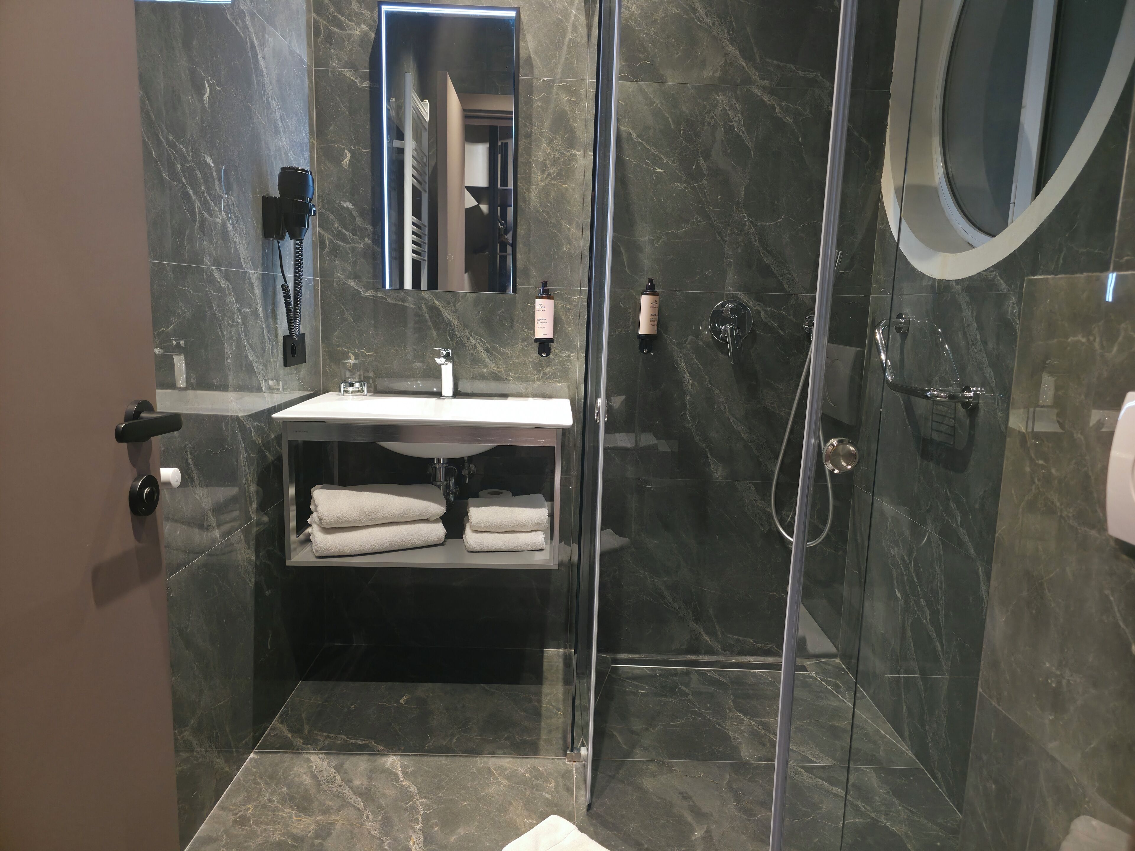 A modern bathroom with a glass-enclosed shower, a marble-tiled wall, a sink with a mirror, and neatly folded towels.