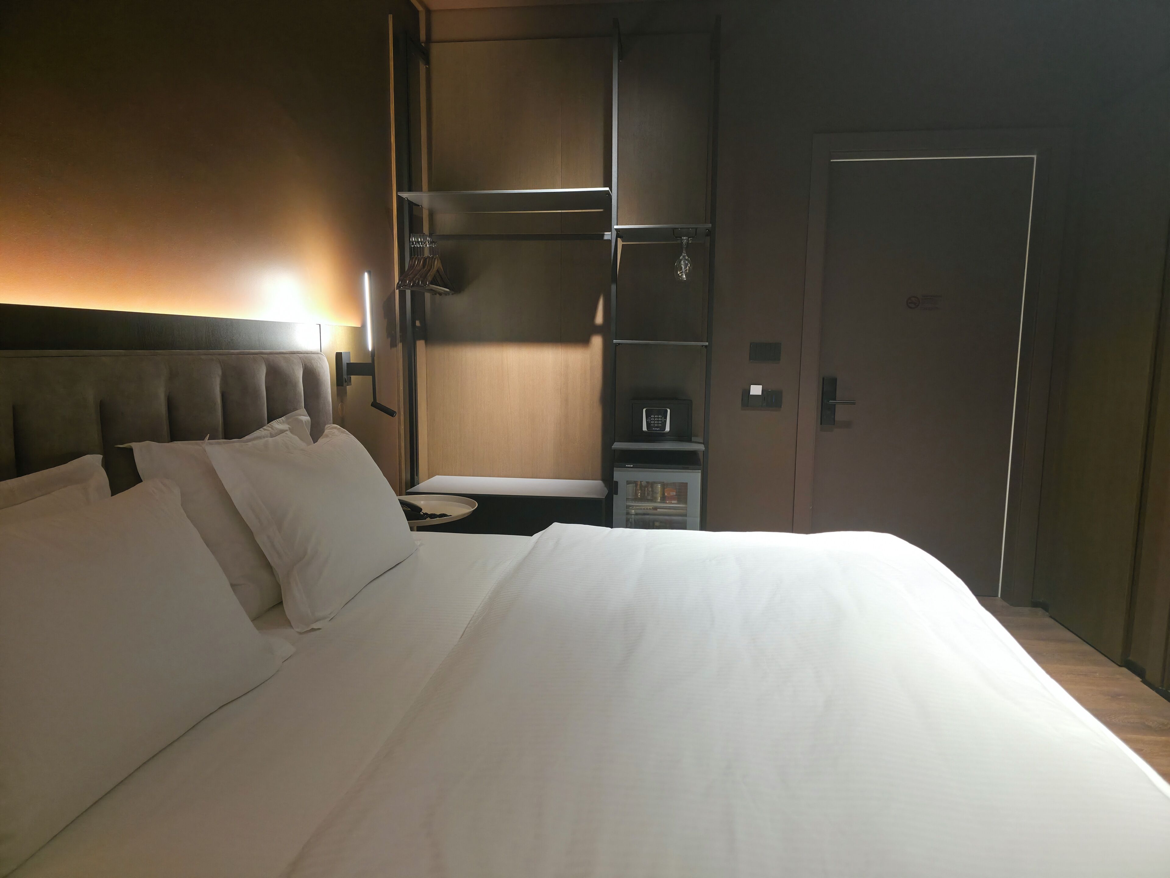 A hotel room with a large bed, a bedside table, a wardrobe, and a minibar.