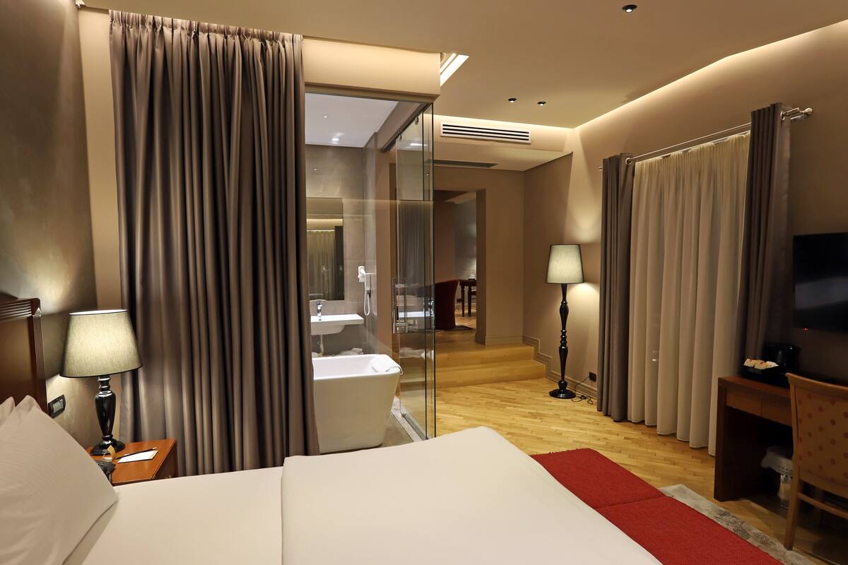 A hotel room with a large bed, a desk, a TV, a bathroom with a bathtub, and a glass partition.
