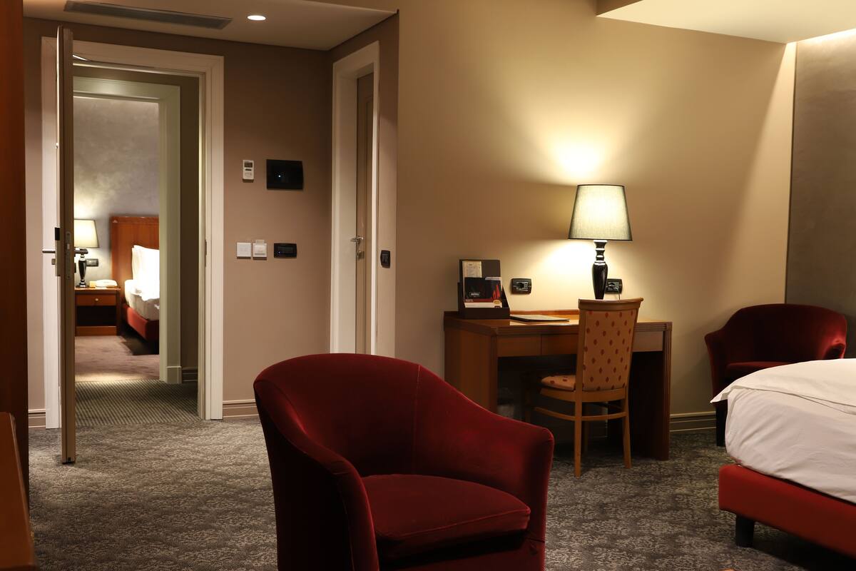 A hotel room with a bed, a desk, a chair, and a lamp. There is a door leading to another room.