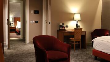 A hotel room with a bed, a desk, a chair, and a lamp. There is a door leading to another room.