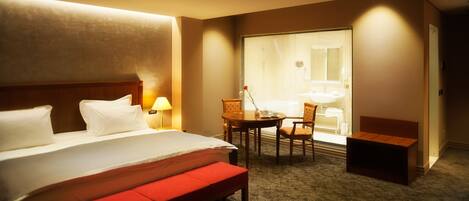 Una habitación de hotel con una cama grande, una mesita con lámpara, una silla y un banco.