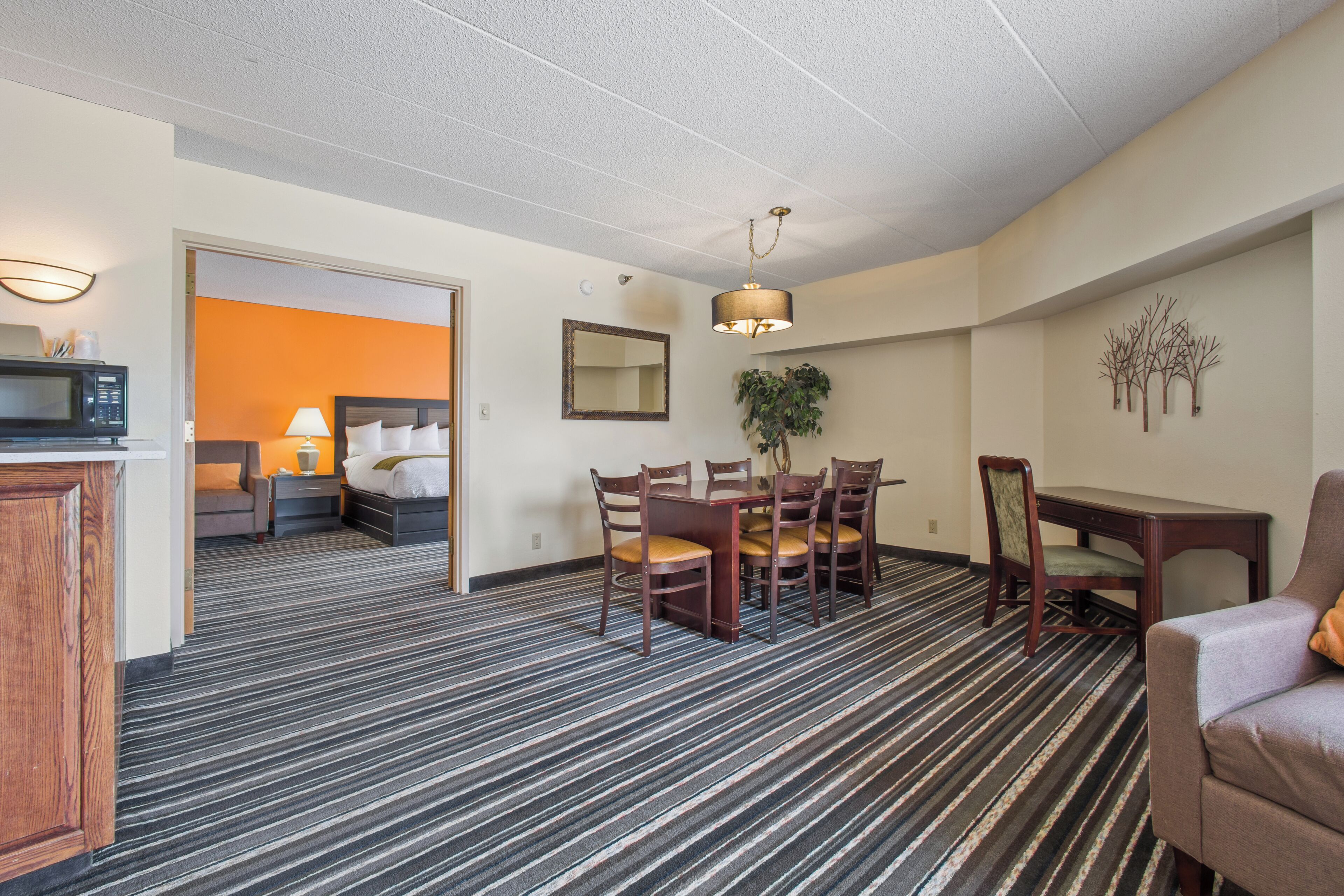 A hotel room with a striped carpet, a dining area with a table and chairs, a microwave, a lamp, and a plant.