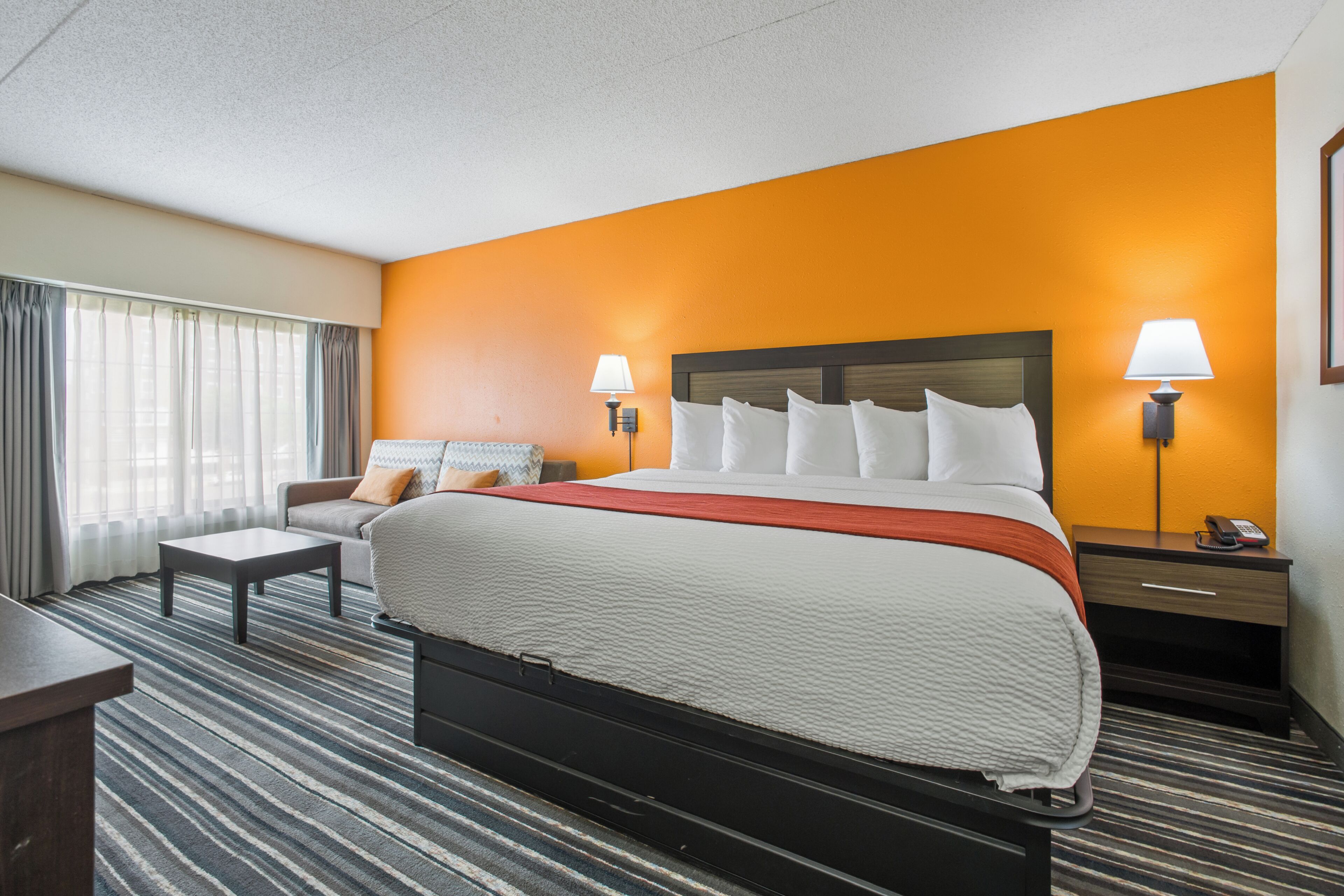 A hotel room with a large bed, a sofa, a desk, and a nightstand with a lamp.