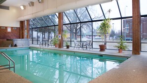 An indoor swimming pool with clear glass walls, potted plants, and a seating area with tables and chairs. - Crossroads Hotel at Huron Event Center (Huron)