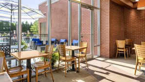 An indoor patio area with wooden tables and chairs, large windows, and a brick wall. - Hyatt Place Memphis/Germantown (Memphis)
