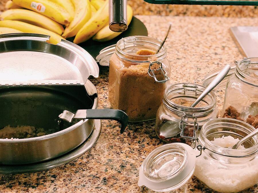 Keittiön työtasolla on metallinen paistinpannu, purkki maapähkinävoita, purkki sokeria, purkki suolaa ja nippu banaaneja.