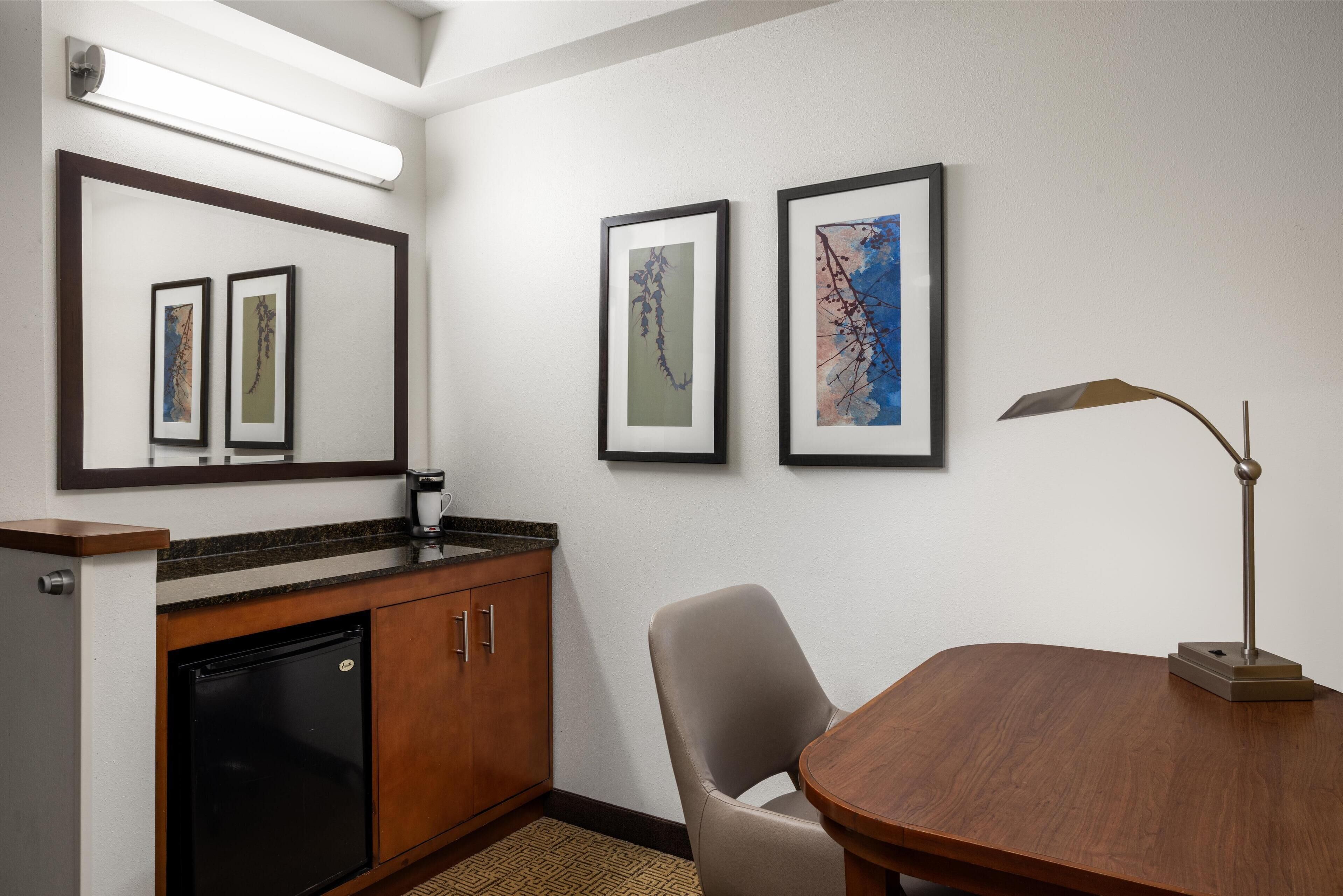 A hotel room with a wooden desk, a chair, a small refrigerator, a coffee maker, and framed artwork on the wall.