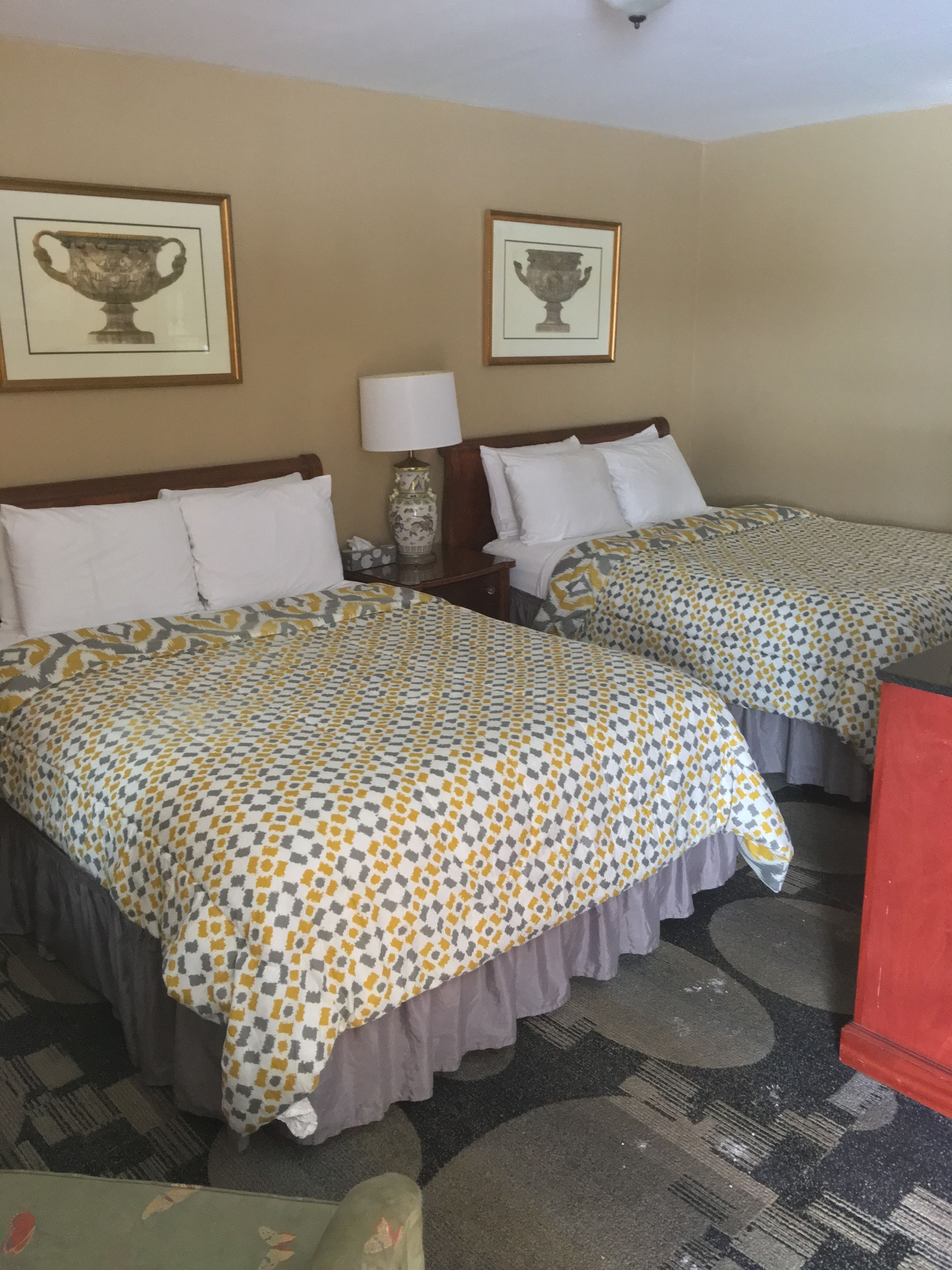 Habitación de hotel con dos camas, una mesita de noche con lámpara y dos cuadros en la pared.